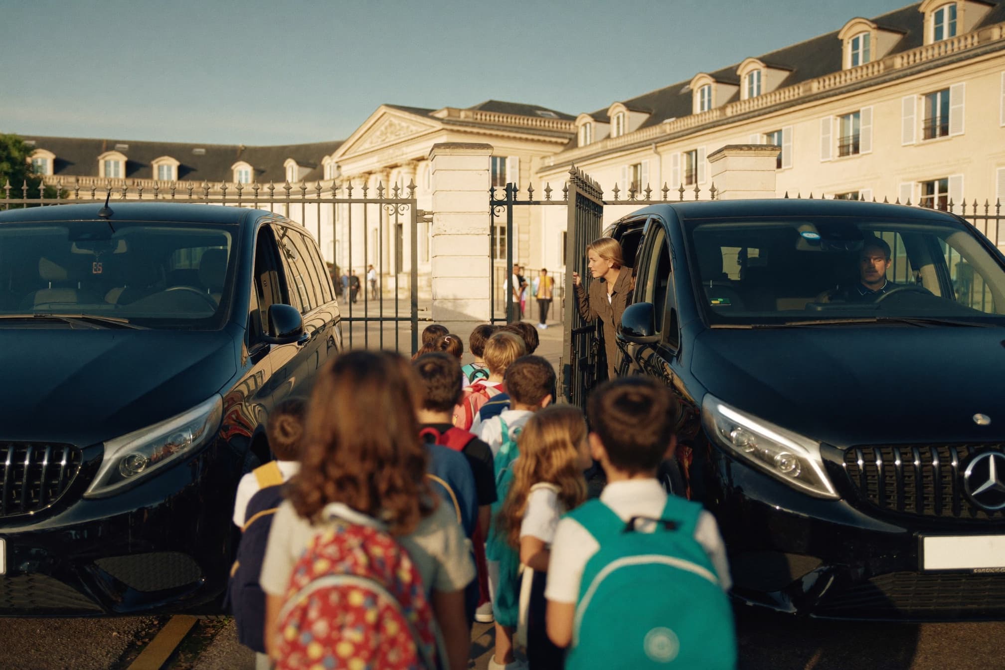 Des élèves descendus de leurs navettes franchissent le portail de l'école, accueillis par une représentante de l'établissement.